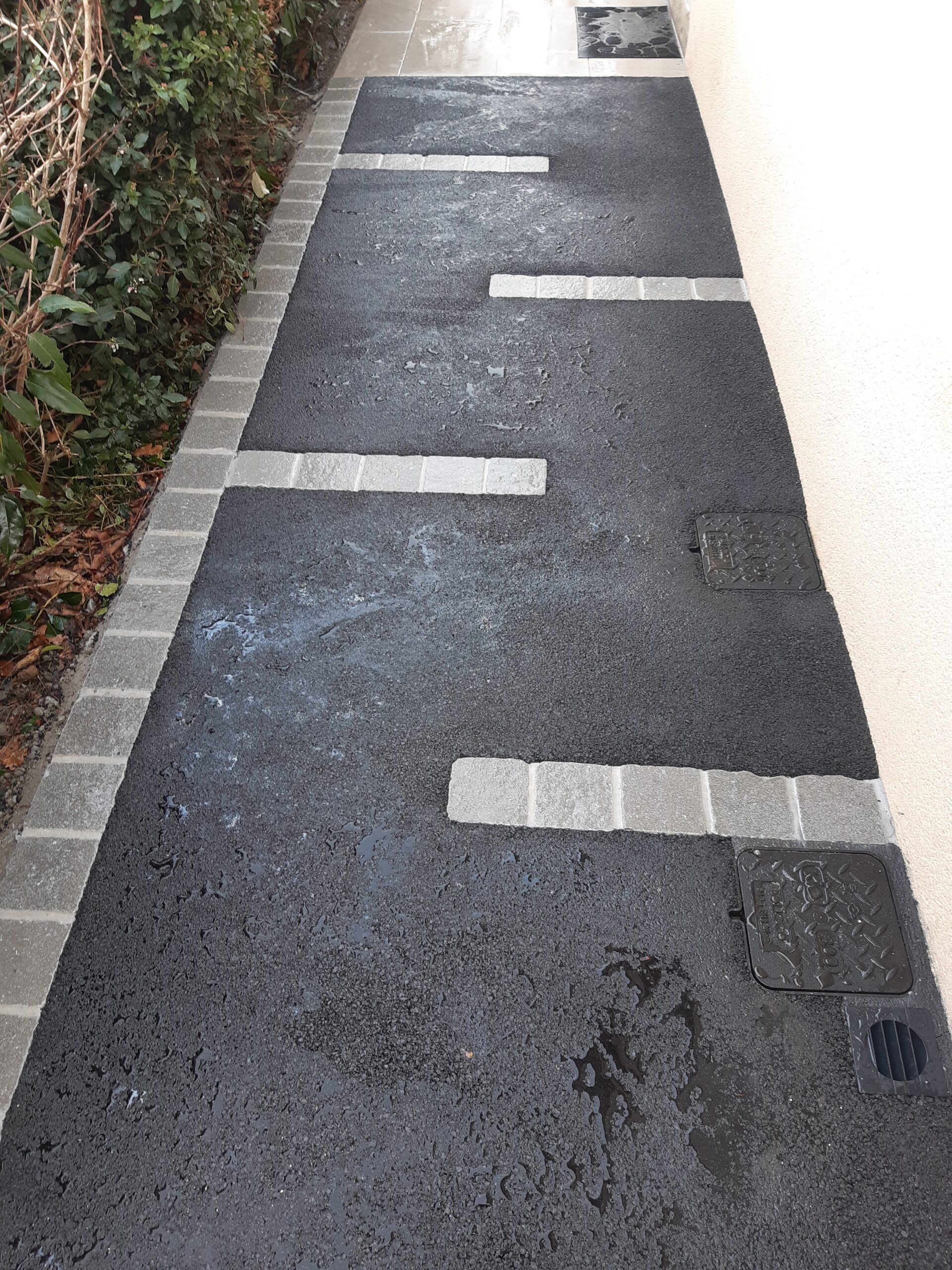 Allée en enrobé noir le long d'une maison, rythmée de bandes de pavés clairs
