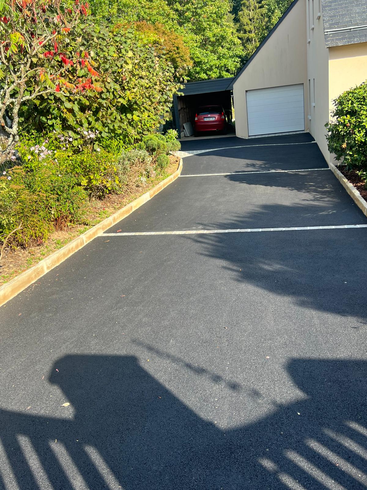Allée en enrobé noir menant à un garage, bordée de massifs